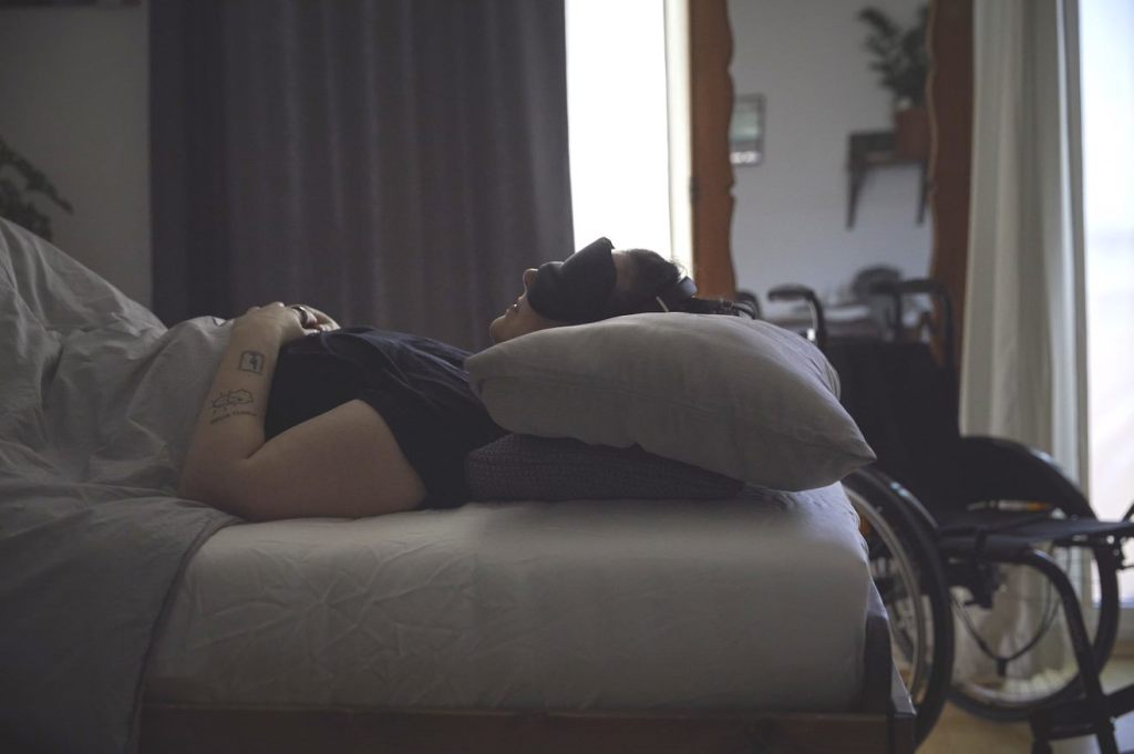 A young white woman lies in bed. She wears an eye mask to block out light. The curtains are drawn but we can see it's daylight outside. There is a wheelchair near the bed.