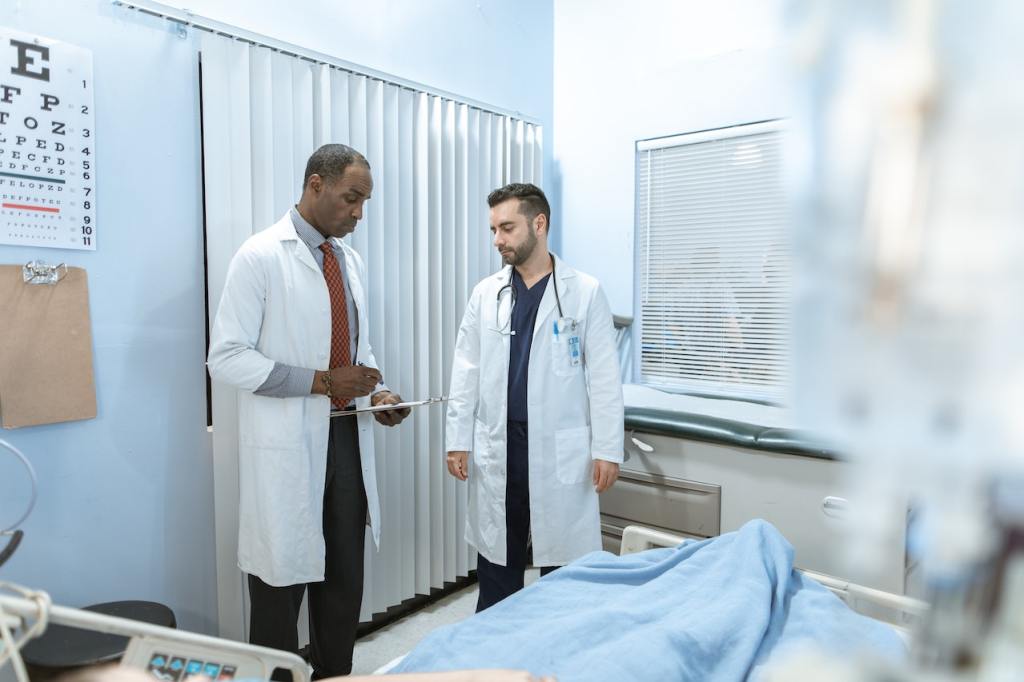 In a hospital patient's room, a middle-aged doctor goes over a patient's chart, while a young doctor looks on.