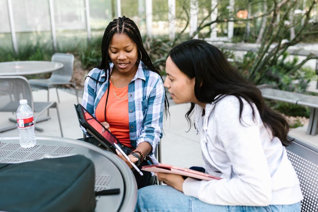 Two young women sit at an outdoor table, one holds a tablet, and both are looking at the tablet with interest.