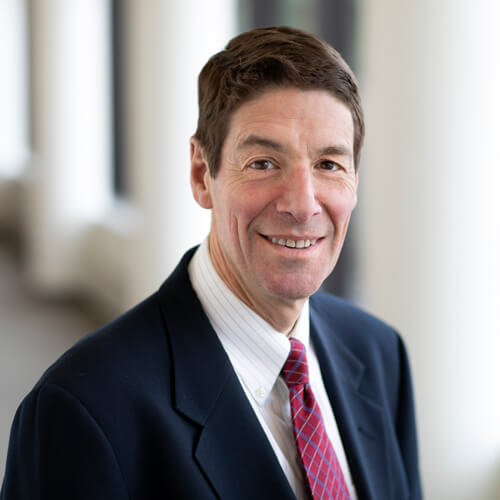 Photo: Dr Howard Andrew Selinger, a white man in his forties wearing a suit and a red tie, smiling.