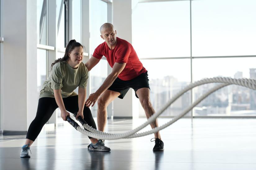 Stock photo: a bright, spacious room. A young white woman who may have Down Syndrome swings two heavy white ropes, smiling widely. A fitness trainer, a tall white man, guides her.
