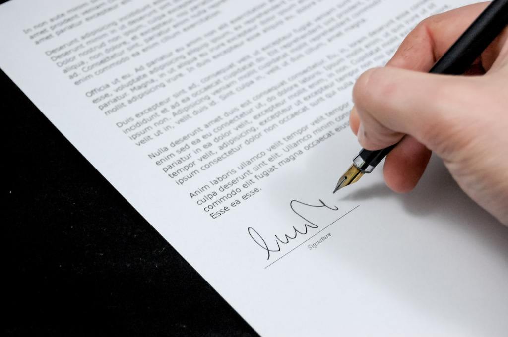 Stock photo: a close-up of a person's hand holding an old-fashioned pen, signing a printed letter. If you look closely, the text of the letter is in Latin.